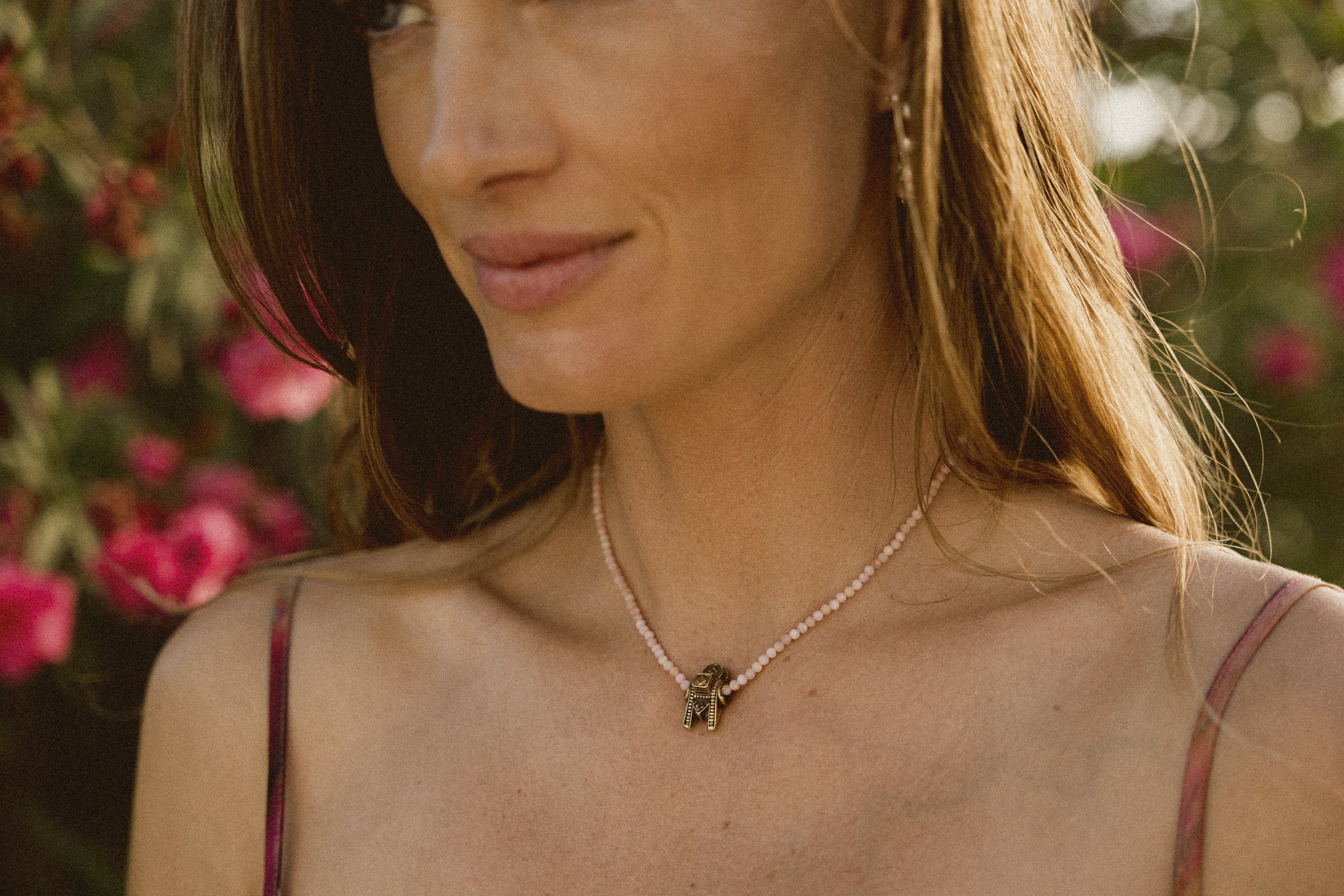 A model wearing a Thali charm on a pink beaded necklace standing against a backdrop of flowers