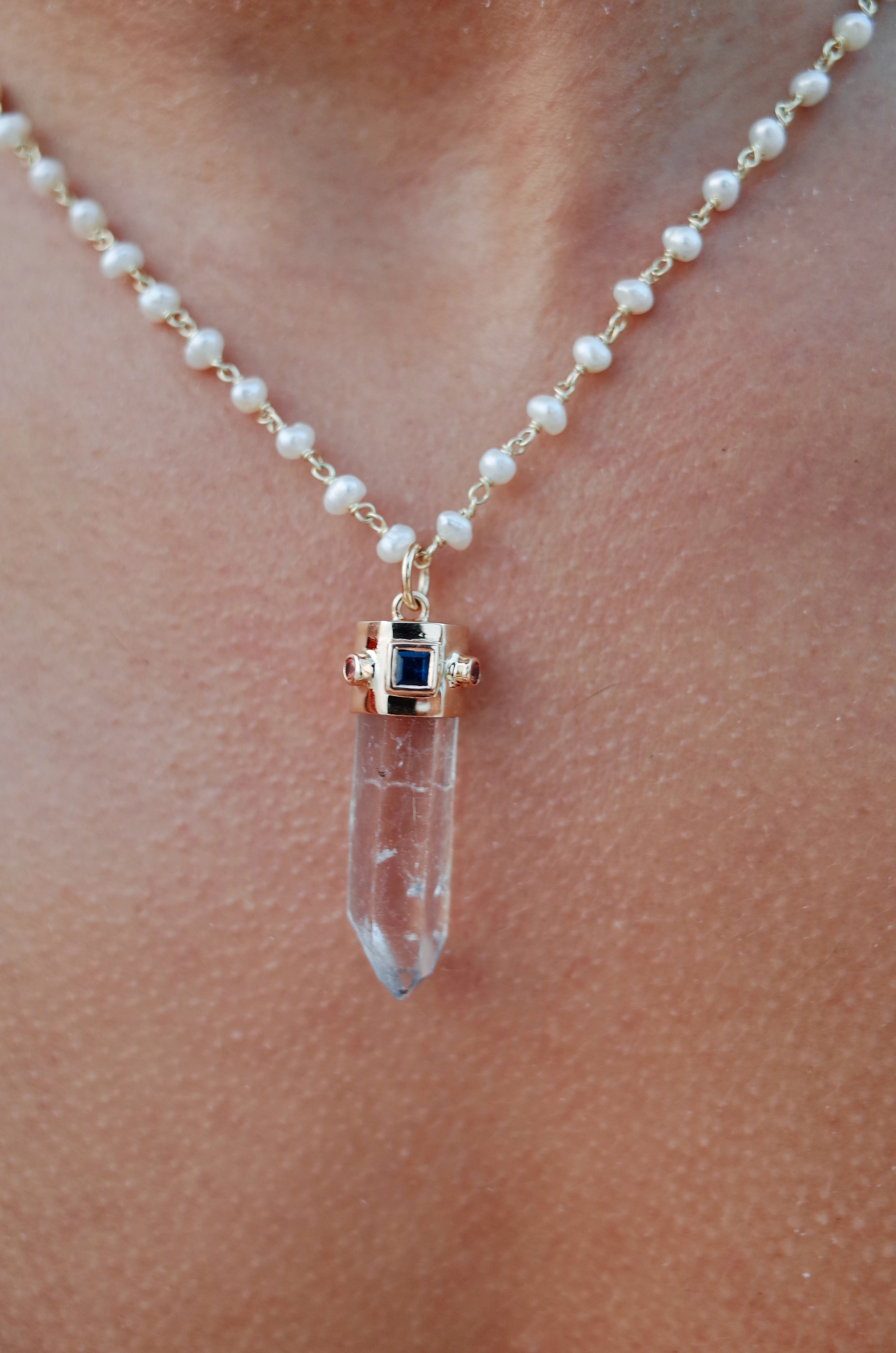 The chest of a model wearing the quartz natural point, blue sapphire and orange sapphire amulet on a pearl beaded necklace