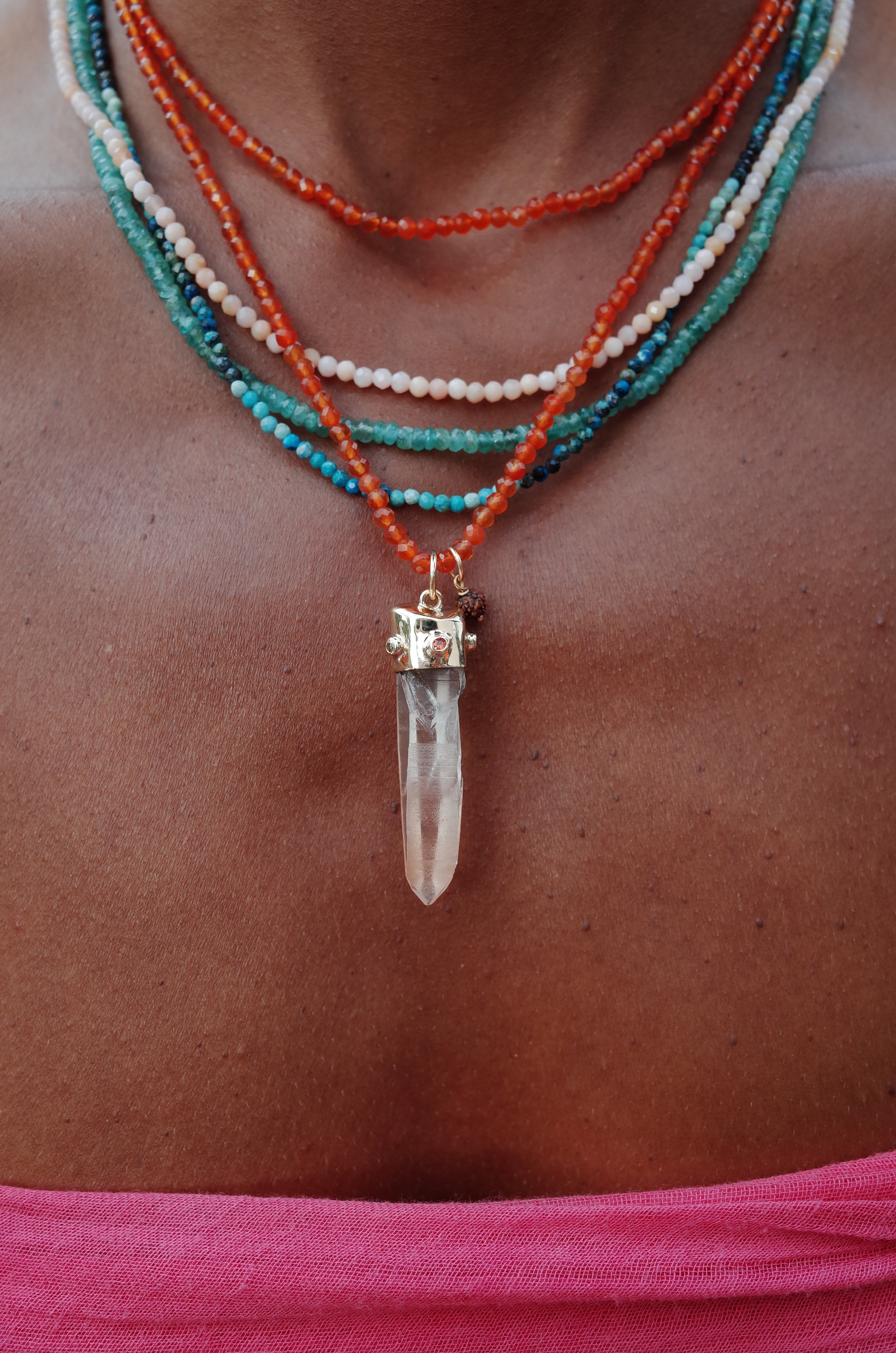 A close-up image of a person wearing a necklace with a large Salmon Lemurian Quartz crystal pendant, accompanied by smaller orange sapphire and white diamond accents, set in 18ct gold. Other colorful beaded necklaces are also visible in the background.