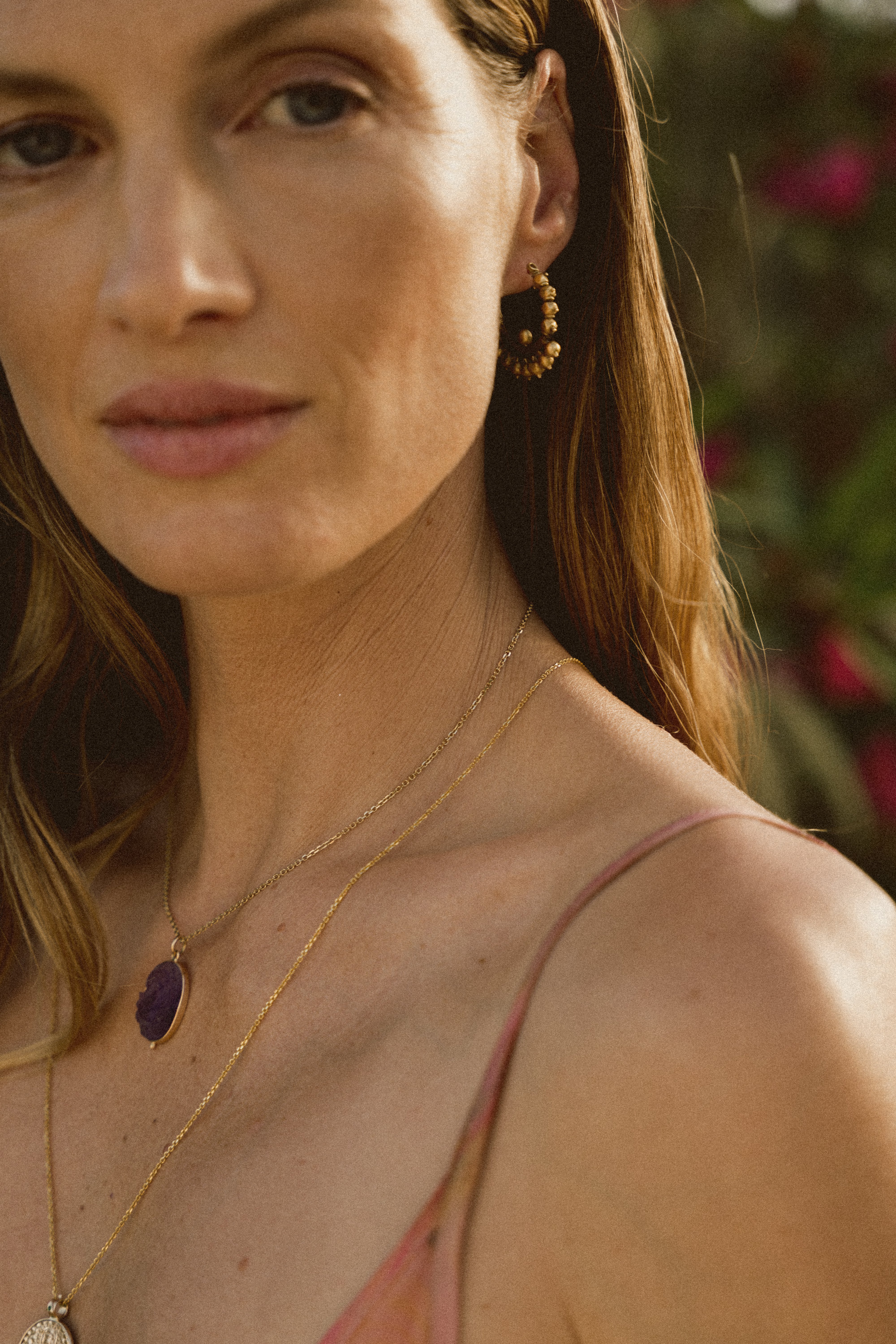 A close up of a model against a backdrop of flowers wearing a gold Nath earring and necklaces. 