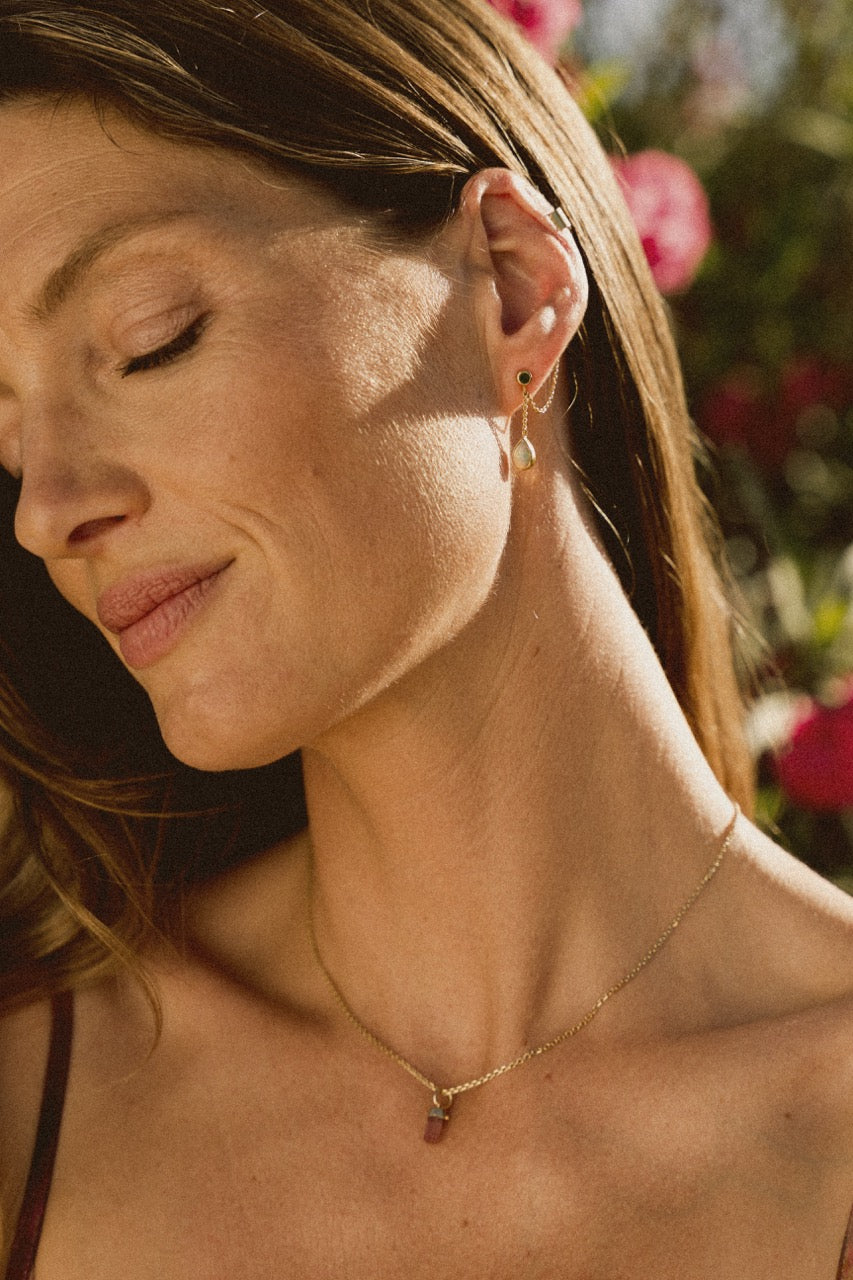 A model wearing a pink tourmaline baby amulet necklace with a gold chain standing against a floral backdrop