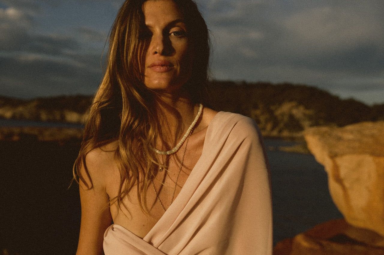 A woman wearing a pink dress and an opal necklace with a gold clasp, standing by a body of water with a rocky background.