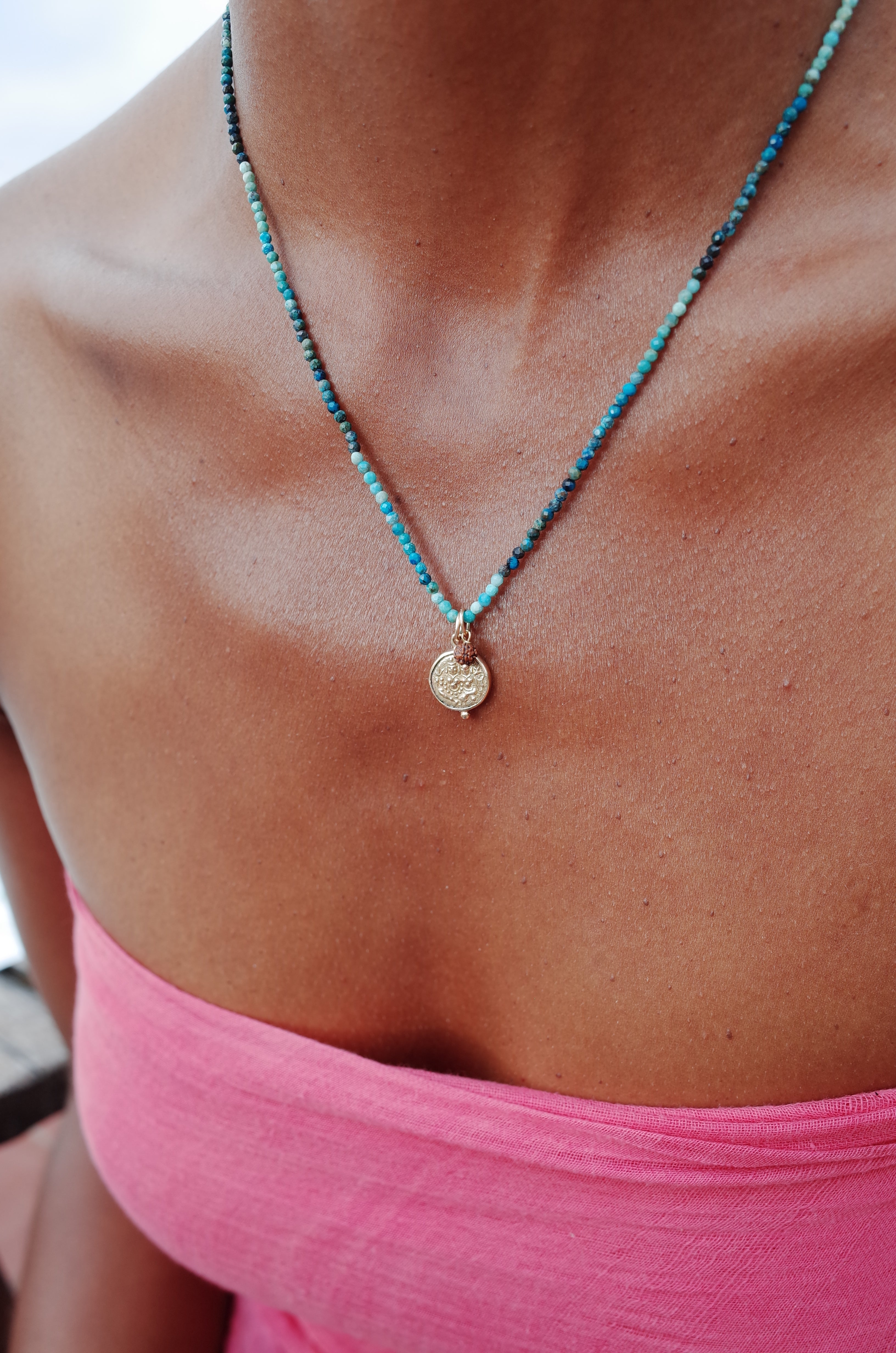 'Union' gold coin necklace worn on string of turquoise beads with rudraksha bead charm on a model wearing a pink top 